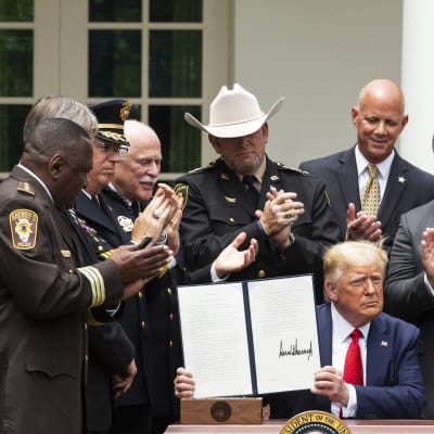 Istuva Trump esittelee allekirjoitustaan. Ympärillä seisoo poliiseja.