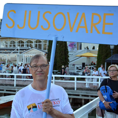 Årets sjusovare 2018, Tapani Bastman, står i Nådendal med en stor blå skylt där det står "Sjusovare"