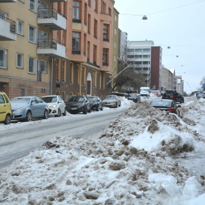 Stora snöhögar på en smal väg, flera bilar står parkerade längs vägen. 