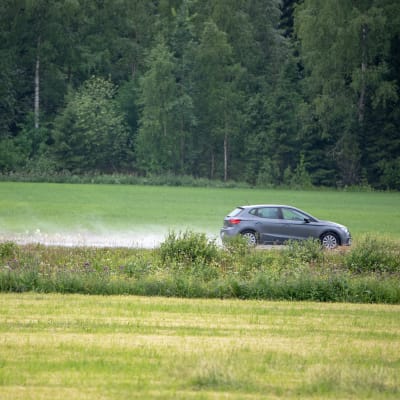 Henkilöauto ajaa sateisella tiellä peltojen keskellä.