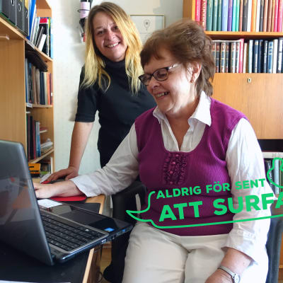 En äldre kvinna sitter framför en bärbar dator. Bakom henne står en kvinna i medelåldern som ler och ser in i kameran. På bilden finns också en logga med texten "Aldrig för sent att surfa".