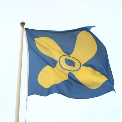 Kimitoöns blåa flagga med en gul propeller i mitten mot en ljusblå himmel.