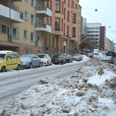 Stora snöhögar på en smal väg, flera bilar står parkerade längs vägen. 