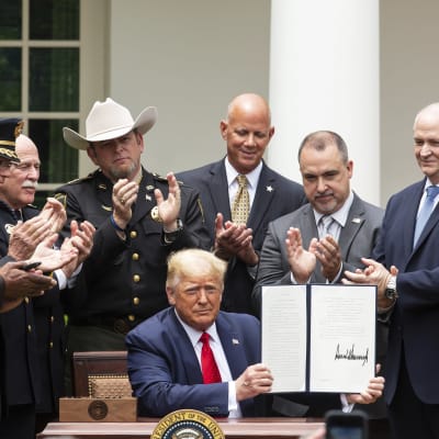 Donald Trump sitter och håller upp ett avtal, runtomkring honom applåderar män i kostym och uniform.