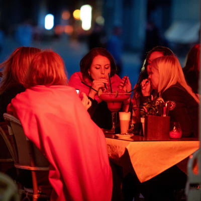 Ett gäng kvinnor sitter på en uteservering på kvällen och dricker en och samma stora dryck ur sugrör. Det lyser ett rött sken på kvinnorna.