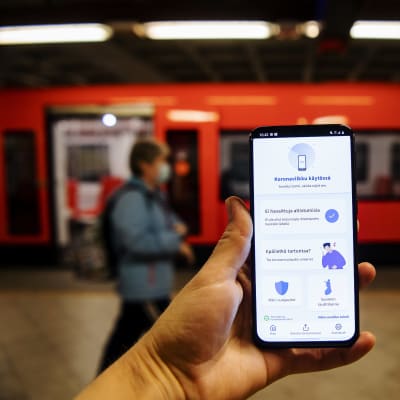 En hand håller upp en telefon med appen Coronablinkern, på en metroperrong.