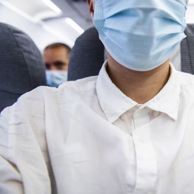 Manlig flygpassagerare i vit skjorta och ljusblått munskydd ombord på ett passagerarplan.