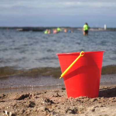 En röd plasthink på en sandstrand med barn som simmar i vattnet i bakgrunden. 
