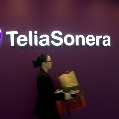 Telia Soneras logotyp mot en vinröd bakgrund. I förgrunden suddigt en kvinna som går förbi.