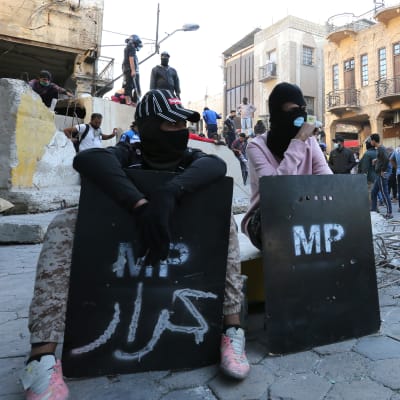 Demonstranter med sköldar sitter på gatan framför en barrikad i Bagdad