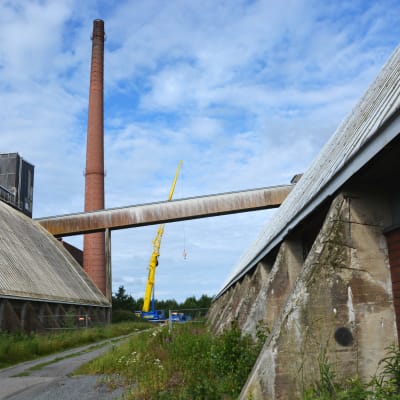 På bilden ser man skorstenen vid sockerfabriken på avstånd. Brevid den står en gul lyftkran. I förgrunden ser man två lagerbyggnader.