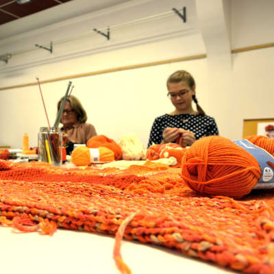 Hannele Palm och Veronica Lönnqvist sitter och stickar i ett klassrum. Framför dem ligger mängder av garn i olika orange nyanser.