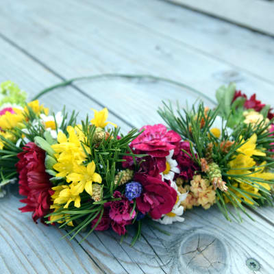 En blomsterkrans som bundits på en grov metalltråd och som bara har dekorerats i en halvcirkel. Växterna som använts ör röd nejlika, gul allium, prästkrage och tallris med späda gröna kottar.