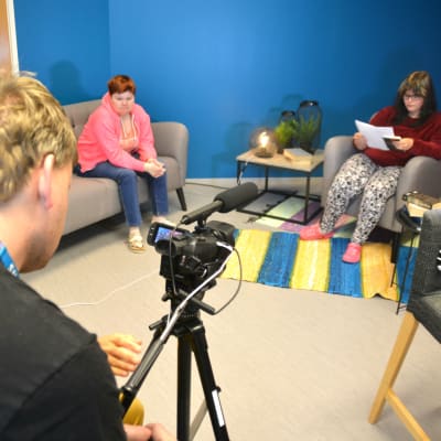Opiskelija kuvaa jalustalla olevalla videokameralla nojatuolissa istuvaa henkilöä.