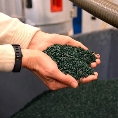 Händer som håller i gröna plastpelletar ovanför ett stort fabrikskärl.