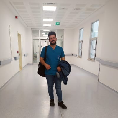 Hoitajan työasuun pukeutunut Ali Dag seisoo sairaalan käytävässä ja katsoo kameraan.