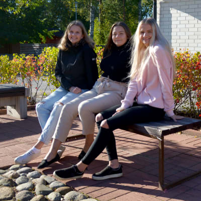 Tre unga kvinnor sitter på en bänk framför en skolbyggnad.