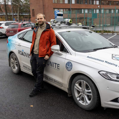 Tutkija Jyri Maanpää Maanmittauslaitoksen Paikkatietokeskuksesta seisoo ARVO-auton vieressä, joka on enimmäkseen valkoinen ja sen kyljissä on erilaisia tekstejä.