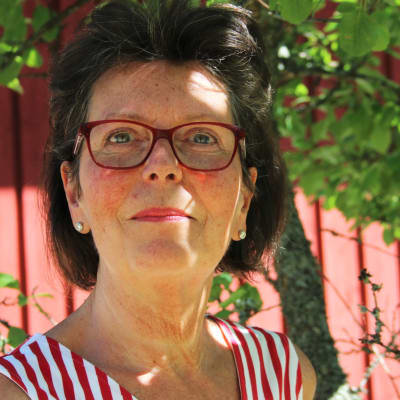 En kvinna med mörkt hår, glasögon med röda bågar och röd-vit (polka) randig topp i en lummig trädgård med rött trästaket, träd och sol