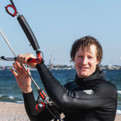 Man i våtdräkt står på en sandstrand i solskenet och håller i en segeltrapets som är fäst runt hans midja med en sele.