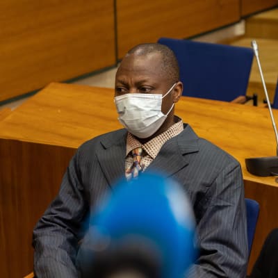 Gibril Massaquoi iklädd kostym i en rättssal. I förgrunden skymtar kameror och mikrofoner.