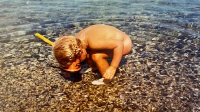 En liten flicka i vattenbrynet med snorkel och grodögon tittar koncentrerat ner mot bottnet. Clara Törvall som barn.
