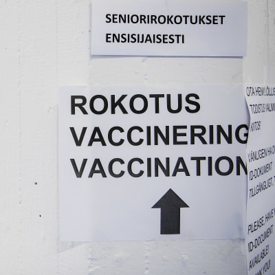 Skylt bland annat med texten vaccinering och till vänster en kvinna som bär munskydd. 85-åringar och äldre samt närvårdare vaccinerades på Busholmen i Helsingfors den 1 februari  2021.