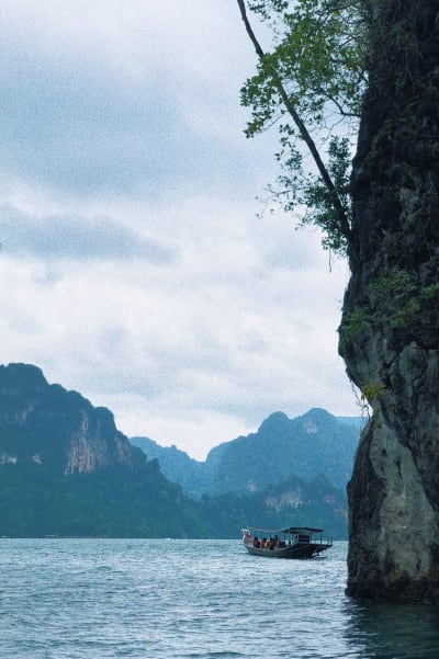 Båt som åker i vatten med klippa i förgrunden och berg och skog i bakgrunden.