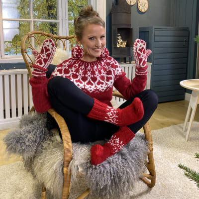 Nainen istuu tuolissa. Stömsövillapaita, lapaset, sukat ja joulukuusenpallot päälle.