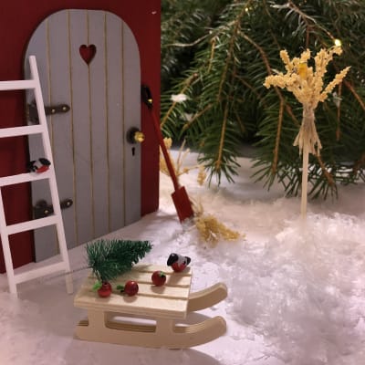 Ett miniatyrlandskap med en tomtenissedörr, snö och småfåglar, en kälke, stege och havrekärvar.