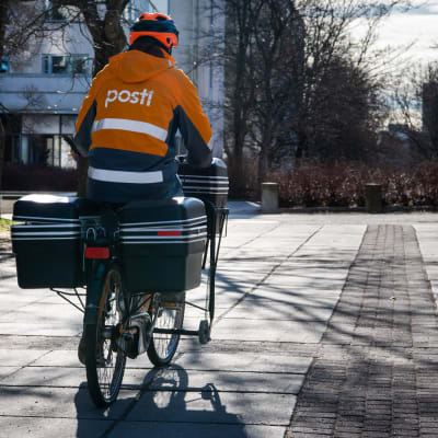 En brevbärare på cykel. Brevbäraren har på sig en orange rock med texten "Posti". Hen har ryggen vänd mot kameran.