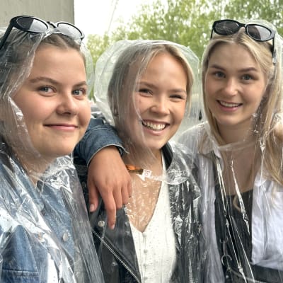 Kolme nuorta naista hymyilee kameralle päällään kertakäyttöiset sadetakit.