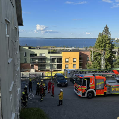 Vaalean kerrostalon edessä seisoo pelastushenkilökuntaa kantaen paloletkuja. Parkkipaikalla on yksi paloauto.