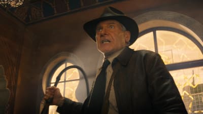 En arg Indiana Jones (Harrison Ford) med läderrock och hatt står med piskan i handen i en stor och hög sal.