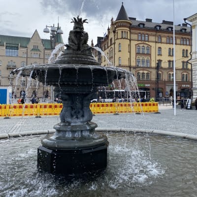 Etualalla on kivinen suihkulähde, jossa vesi virtaa. Taustalla näkyy Tampereen keskustan korkeita kivirakennuksia ja kelta-punainen työmaa-aita.