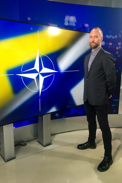 Man står framför en skärm med grafik av Natos logo samt Sveriges och Finlands flaggor.