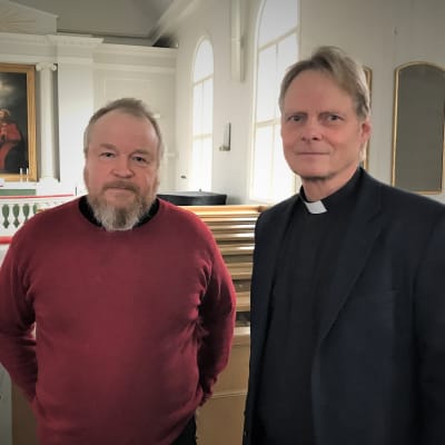 Kaksi miestä seisoo kirkon käytävällä. Oikean puoleinen on mustiin pukeutunut yhtymäjohtaja, vasemmalla punaiseen paitaan pukeutunut pappi. . 