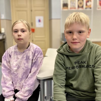 Kaksi ala-asteikäistä lasta istuu koululuokassa ja katsoo kameraan. Vasemmalla istuvalla tytöllä on yllään lilan sävyinen pitkähihainen paita, oikealla istuvalla pojalla maastonvihreä huppari.