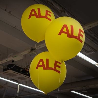Tre gula heliumballonger som det står ALE på. De finns inomhus i en affär och affärens tak syns i bakgrunden.