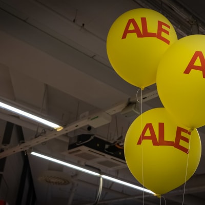 Tre gula heliumballonger som det står ALE på. De finns inomhus i en affär och affärens tak syns i bakgrunden.