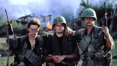 Kolme nuorta jalkaväensotilasta sotatantereella, likaisissa univormuissaan aseet olalla, pitävät käsiään toistensa hartioilla.