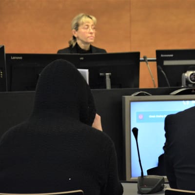 Den åtalade kvinnan fotograferad bakifrån med en uppdragen huva. Bredvid sin man. I bakgrunden syns den kvinnliga tingsrättsdomaren.