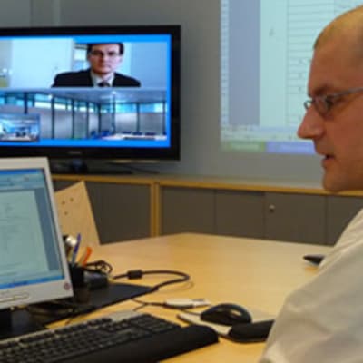 Kuvassa mies tietokonepäätteen ääressä taustalla televisioruudulla videoneuvotteluyhteydessä toinen mies.