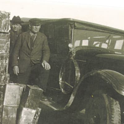 Kuvassa pirtukanistereita, auto ja kaksi miestä. Takavarikoitu spriilasti odottaa jatkokuljetusta vuonna 1930.