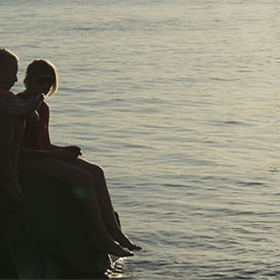 Kaksi tyttöä istuu kivellä vedenäärellä auringonlaskussa.
