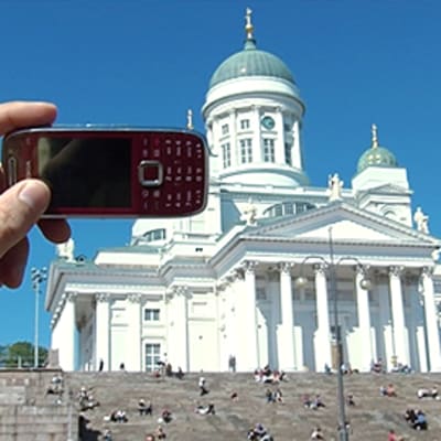 Miehen käsi pitää matkapuhelinta ottaakseen valokuvan Helsingin Tuomiokirkosta.
