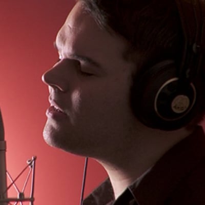 Mikael Konttinen laulaa mikrofoniin studiossa kuulokkeet korvillaan ja silmät kiinni. Taustallaan punertava kajo.