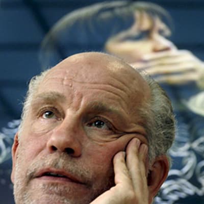 Näyttelijä John Malkovich pitää kämmentä poskellaan näytelmänsä Infernal Comedy esittelytilaisuudessa. Taustalla näytelmän mainosjuliste.