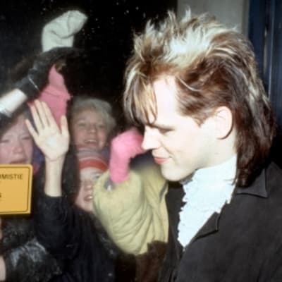 Dingo-yhtyeen laulusolisti Neumann (Pertti Nieminen) nuorten itsenäisyyspäiväjuhlassa 1985. Neumann seisoo ovella, jonka takana huutavia nuoria tyttöjä. Dingon ihailijoita lasioven takana. 