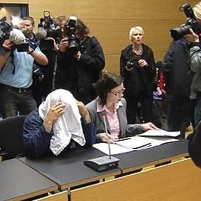 Kuvaajat piirittävät T-paidalla kasvojaan peittävää syytettyä Helsingin käräjäoikeudessa.
